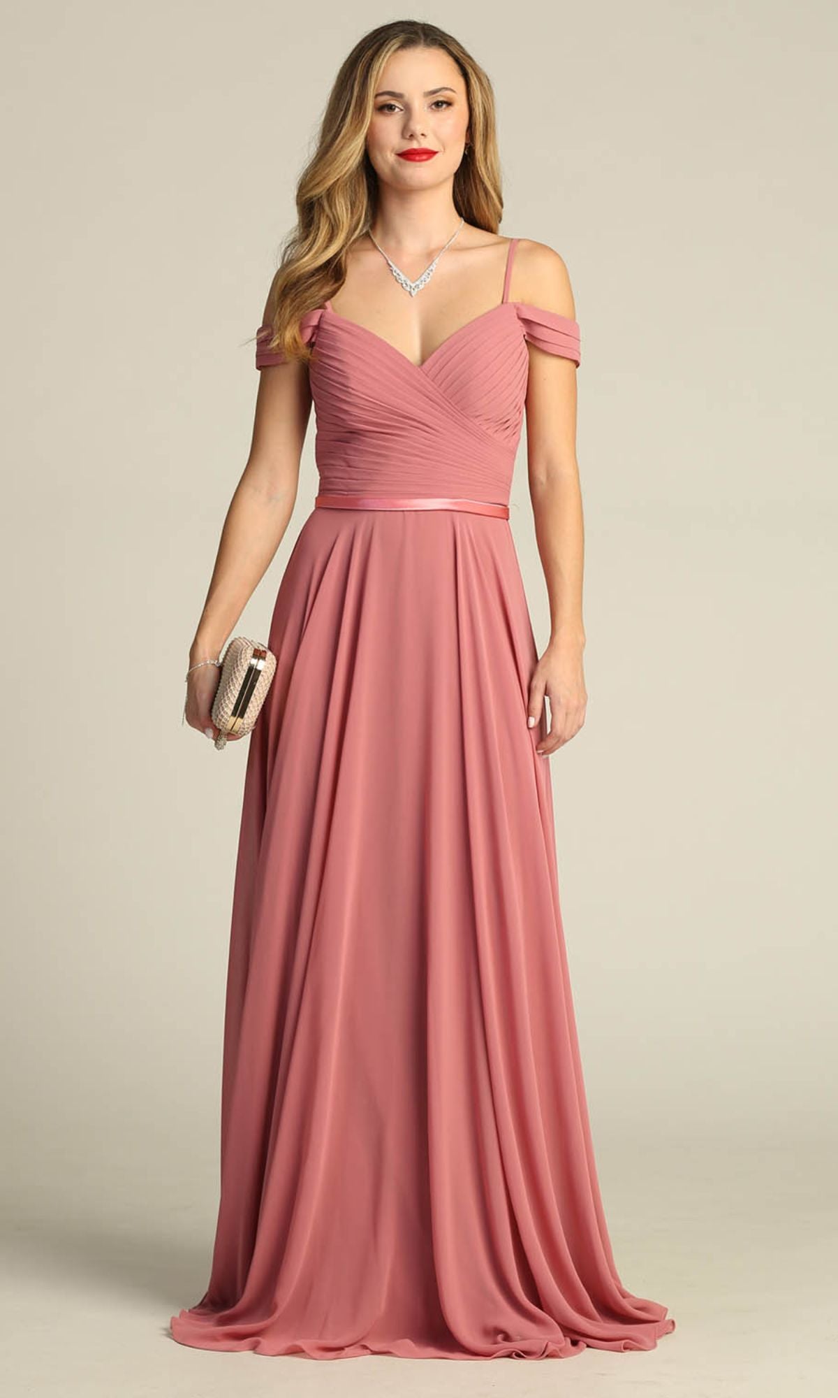 A woman in the Chicas C8997 long A-line off-shoulder prom dress in dusty rose holds a beige clutch and wears a necklace, standing against a plain light background.